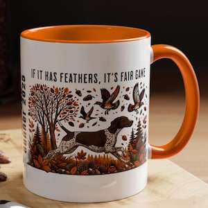 May include: White ceramic mug with an orange interior and handle. The mug features a hunting scene with a dog, birds, and trees, with the text "IF IT HAS FEATHERS, IT'S FAIR GAME". A spoon and a pastry are in the foreground.