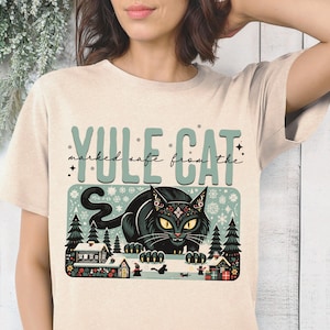 May include: A beige t-shirt with a graphic print of a black cat with a snowflake design on its head. The cat is sitting in front of a snowy winter scene with houses and trees. The text "YULE CAT marked safe from the" is printed above the cat.