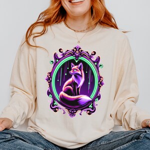 May include: A cream colored sweatshirt with a purple and green ornate frame featuring a purple fox with a green glow. The fox is looking up at a starry sky.