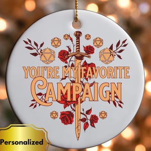 May include: A white ceramic ornament with the text "YOU'RE MY FAVORITE CAMPAIGN" in orange. A sword and red roses are in the center. The ornament has a gold string for hanging and a gold "Personalized" sticker.