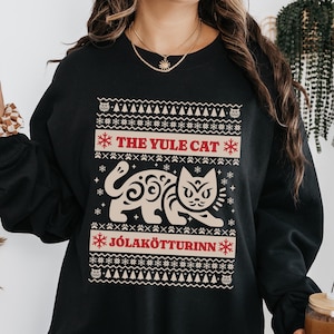May include: Black sweatshirt featuring a festive design with the text "THE YULE CAT" and "JÓLAKÖTTURINN" in red. The design includes a stylized cat illustration and snowflake accents, perfect for holiday wear.