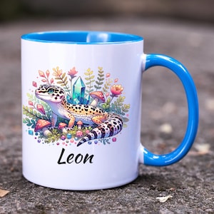 Puede incluir: Taza de cerámica blanca con interior y asa azules. La taza presenta una colorida ilustración de un gecko con setas, cristales y flores. El nombre "Leon" está impreso debajo.