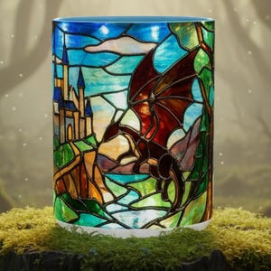 Op de afbeelding: Cilindrische glas-in-lood lamp met een draak die over een kasteel vliegt. Het kunstwerk gebruikt een palet van blauw, groen, bruin en rood. De lamp staat op een mosachtig oppervlak, wat een fantasiethema creëert.