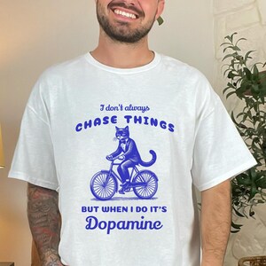 May include: White t-shirt with a blue graphic of a cat in a suit riding a bicycle. The text on the shirt reads "I don't always chase things but when I do it's Dopamine."