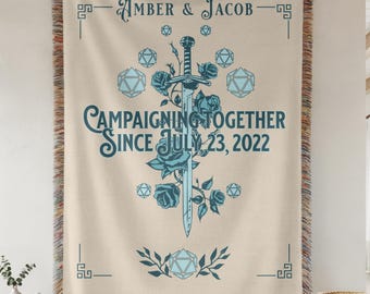 Può includere: Coperta in tessuto color crema con un design decorativo. La coperta presenta i nomi "Amber & Jacob" e la frase "Campaigning Together Since July 23, 2022" in verde acqua. Sono inclusi anche una spada, rose e forme geometriche.
