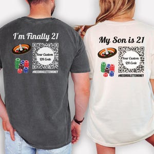 May include: Two t-shirts, one dark gray and one off-white, with casino-themed graphics. The dark gray shirt says "I'm Finally 21" and the off-white shirt says "My Son is 21". Both shirts have a roulette wheel, stacks of chips, and a QR code.