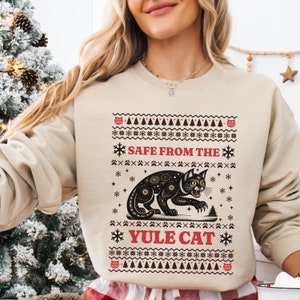 Peut inclure: Un sweat-shirt beige avec un motif festif représentant un chat noir à motif étoilé. Le texte "SAFE FROM THE YULE CAT" est imprimé en rouge sur le sweat-shirt.