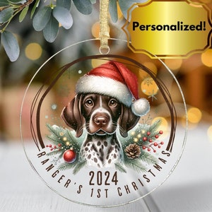 May include: Clear, round Christmas ornament featuring a German Shorthaired Pointer wearing a Santa hat. The ornament includes the text "Ranger's 1st Christmas 2024" and is decorated with festive greenery and a red ornament.