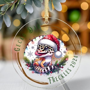 Peut inclure: Une boule de Noël ronde en verre avec une bordure brune et un fond blanc. La boule représente un gecko de dessin animé portant un bonnet de Père Noël et entouré de houx, de flocons de neige et de lumières de Noël. Le texte "Gecko Dad Freckles 2024" est écrit autour du gecko.