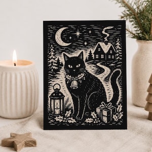 May include: A black and white greeting card featuring a linocut-style illustration of a black cat wearing a bell collar, sitting beside a lantern and a gift box in front of a house, pine trees, a crescent moon, and stars in the night sky.