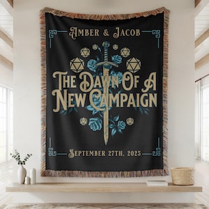 May include: Black tapestry throw blanket featuring the text "AMBER & JACOB" and "THE DAWN OF A NEW CAMPAIGN". The design includes a sword, blue roses, and dice. The date "SEPTEMBER 27TH, 2025" is also displayed. The blanket has a fringed edge.
