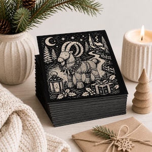 May include: A stack of square greeting cards featuring a black and white linocut-style illustration of a straw goat adorned with a wreath, standing near a lantern, a gift box, and a winter village scene under a crescent moon and stars.