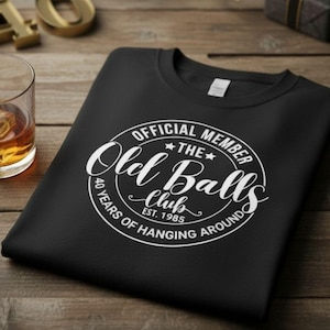 Könnte beinhalten: Schwarzes T-Shirt mit Rundhalsausschnitt und weißem, kreisförmigem Aufdruck. Der Aufdruck lautet "OFFICIAL MEMBER THE Old Balls Club" mit "40 YEARS OF HANGING AROUND" und "EST. 1985" in weißer Schrift. Das Shirt liegt auf einer Holzoberfläche.