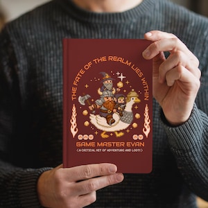 May include: A burgundy notebook with pixelated fantasy art. The cover features a wizard, dwarf, and hooded figure riding a goose, with text that reads "The Fate of the Realm Lies Within" and "Game Master Evan."