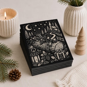 May include: A stack of rectangular greeting cards featuring a black and white linocut-style illustration of a winter scene with a yule log, a lantern, a gift box, a crescent moon, stars, pine trees, and a small house on a snowy hill.