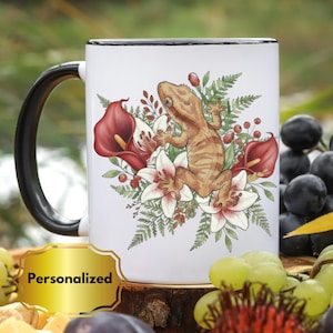 Peut inclure: Mug en céramique blanche avec une anse et un bord noirs. La tasse présente une illustration détaillée d'un gecko entouré de callas rouges, de lys blancs et de feuillage vert. Le mot "Personnalisé" est sur une étiquette dorée.