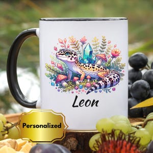 Puede incluir: Taza de cerámica blanca con asa y borde negros, con una colorida ilustración de un gecko, cristales y setas. El nombre "Leon" está impreso debajo. Una pegatina dorada "Personalizado" está en la parte inferior.