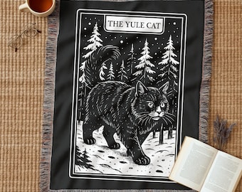Może przedstawiać: Czarny koc z białą, obramowaną ilustracją czarnego kota spacerującego po zaśnieżonym lesie. U góry widnieje napis "THE YULE CAT". Koc ma frędzelkową krawędź.