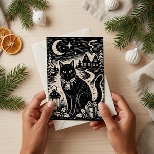 May include: A rectangular greeting card featuring a black and white linocut-style illustration of a black cat wearing a bell collar, sitting near a lantern, a gift box, and holly, with a crescent moon, stars, and a house in the background.