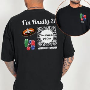 Può includere: T-shirt nero con la scritta "I'm Finally 21" e una grafica di roulette. Include un codice QR con il testo "Your Custom QR Code" e l'hashtag "#NEEDROULETTEMONEY". Una piccola grafica di fiches da poker è presente sul davanti.