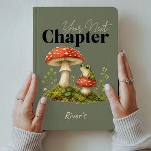 Puede incluir: Un diario de tapa dura color verde salvia con el texto Your Next Chapter en letra negra. La portada muestra una seta grande roja y blanca, una seta más pequeña con una rana verde encima y el nombre River's en una tipografía clara.