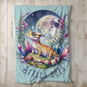 May include: A light blue blanket featuring a gecko illustration with a full moon, crystals, and floral elements. The gecko is yellow, orange, and red. The blanket has the text "Stella 2025" in a script font.