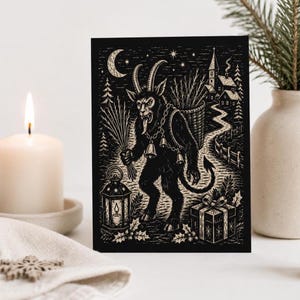 May include: A rectangular greeting card featuring a black and white woodcut-style illustration of Krampus. The figure has horns, a tail, and bells, holding a bundle of sticks near a lantern, a gift box, and a village scene under a crescent moon.