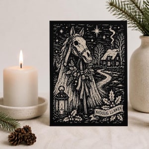 May include: A rectangular greeting card featuring a black and white linocut-style illustration of a horse skull adorned with holly and ribbons. A lantern, a cottage, and a star-filled sky are depicted. The text NADOLIG LLAWEN is on a banner.