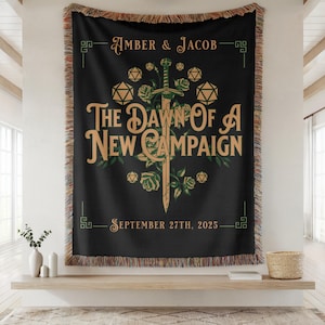 May include: Black throw blanket with a decorative design featuring a sword, roses, and dice. Text reads "AMBER & JACOB" and "THE DAWN OF A NEW CAMPAIGN SEPTEMBER 27TH, 2025". The blanket has a fringed edge.