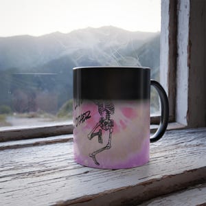 May include: Black ceramic mug with a skeleton graphic and the words "LOVE OTHER". The mug has a black handle and a pink and white background. Steam rises from the mug, suggesting it contains a hot beverage. The mug is placed on a wooden windowsill.