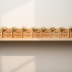May include: A row of wooden memorial blocks with the inscription "In Loving Memory of Maria Terra" and dates. Each block is adorned with a small bouquet of white flowers. The blocks are displayed on a light-colored wooden shelf.