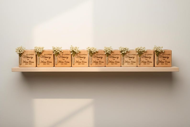 May include: A row of wooden memorial blocks with the inscription "In Loving Memory of Maria Terra" and dates. Each block is adorned with a small bouquet of white flowers. The blocks are displayed on a light-colored wooden shelf.