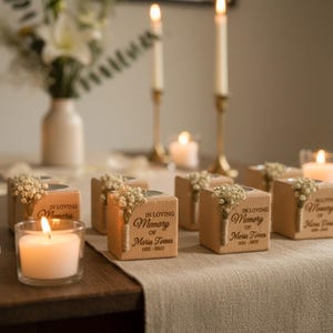 May include: A memorial table setting with lit candles, votives, and floral arrangements. Wooden blocks with the inscription "In Loving Memory" are adorned with small flowers. Tall candlesticks and a vase of flowers are in the background.
