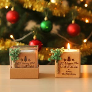 May include: Two wooden Christmas candle holders, one lit, with the text "Merry Christmas" and a small tree design. The holders are decorated with green sprigs and sit in front of a decorated Christmas tree with red and green ornaments.