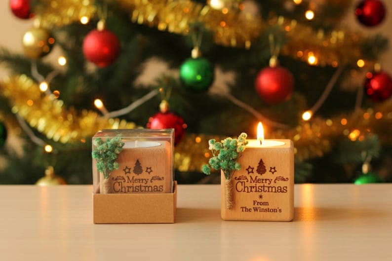 May include: Two wooden Christmas candle holders, one lit, with the text "Merry Christmas" and a small tree design. The holders are decorated with green sprigs and sit in front of a decorated Christmas tree with red and green ornaments.