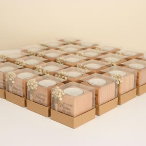 May include: Arrangement of square wooden candle holders with clear plastic tops, each set in a small brown box. The candle holders have a small white candle inside and are decorated with small white flowers. Text is visible on the front.
