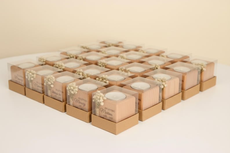 May include: Arrangement of square wooden candle holders with clear plastic tops, each set in a small brown box. The candle holders have a small white candle inside and are decorated with small white flowers. Text is visible on the front.