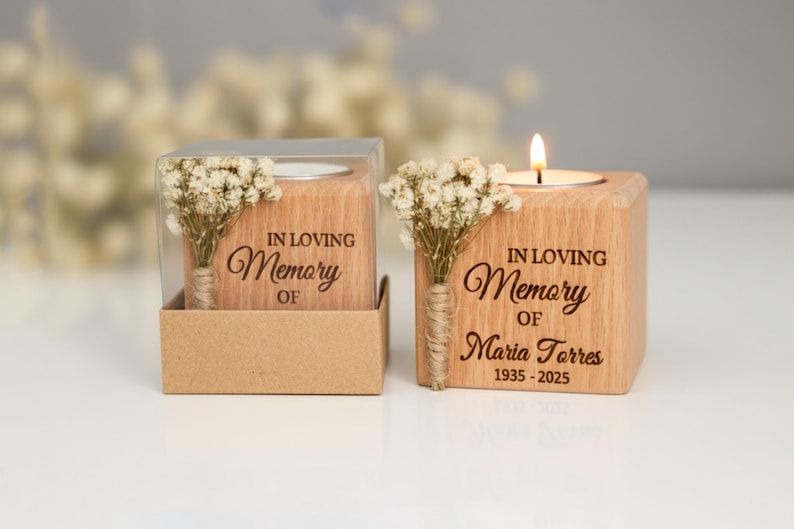 May include: Two wooden memorial candle holders with the text "IN LOVING Memory OF" and a name and dates. One candle is lit. Each holder has a small bouquet of dried flowers.
