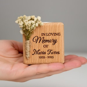 May include: A wooden memorial candle holder with the inscription "IN LOVING Memory OF Maria Torres 1935-2025." It is adorned with a small bouquet of dried white flowers and held in a hand.