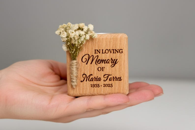 May include: A wooden memorial candle holder with the inscription "IN LOVING Memory OF Maria Torres 1935-2025." It is adorned with a small bouquet of dried white flowers and held in a hand.