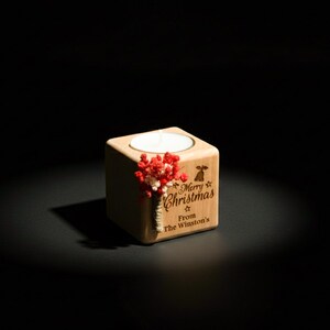 May include: A wooden cube candle holder with a lit white candle. The holder is engraved with "Merry Christmas From The Winston's" and decorated with red and white dried flowers. The background is black.