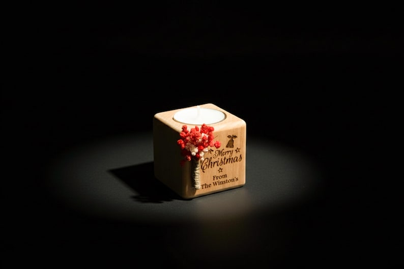 May include: A wooden cube candle holder with a lit white candle. The holder is engraved with "Merry Christmas From The Winston's" and decorated with red and white dried flowers. The background is black.