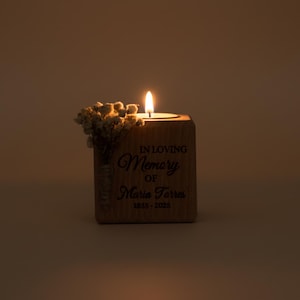 May include: A lit candle in a wooden block with the inscription "IN LOVING MEMORY OF Maria Torres 1935-2025." Dried flowers are attached to the side of the block. The candle flame is bright against a warm, neutral background.