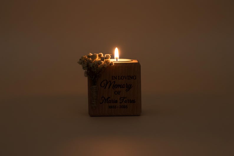 May include: A lit candle in a wooden block with the inscription "IN LOVING MEMORY OF Maria Torres 1935-2025." Dried flowers are attached to the side of the block. The candle flame is bright against a warm, neutral background.