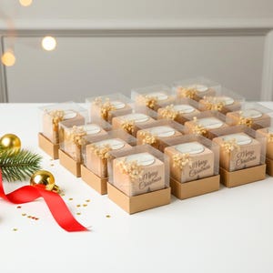May include: Arrangement of Christmas-themed gift boxes. Each box is square, with a clear top, and contains a candle and dried flowers. The boxes are arranged on a white surface, with a red ribbon, gold ornaments, and greenery.