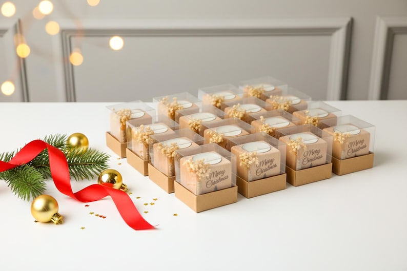 May include: Arrangement of Christmas-themed gift boxes. Each box is square, with a clear top, and contains a candle and dried flowers. The boxes are arranged on a white surface, with a red ribbon, gold ornaments, and greenery.