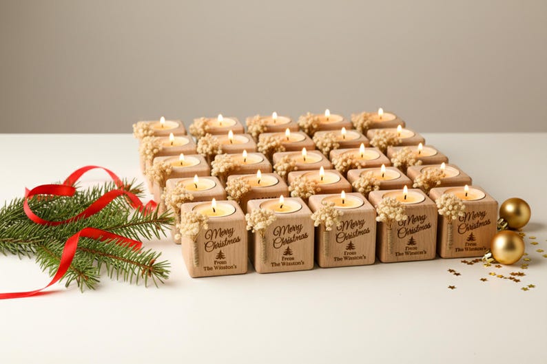 May include: A collection of lit tea light candles in small wooden blocks, arranged in a grid. Each block is decorated with a floral accent and the words "Merry Christmas". A sprig of evergreen and red ribbon are on the left, with gold ornaments.
