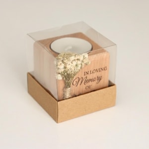May include: A wooden candle holder with a tea light inside, presented in a clear plastic box and a brown cardboard base. The holder is engraved with the words "IN LOVING Memory OF" and features a small bouquet of dried flowers.
