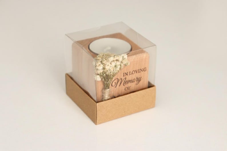 May include: A wooden candle holder with a tea light inside, presented in a clear plastic box and a brown cardboard base. The holder is engraved with the words "IN LOVING Memory OF" and features a small bouquet of dried flowers.