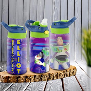 May include: Three water bottles with blue lids, green accents, and colourful designs. One bottle has the name "ELLIOT" and the text "GALACTIC GUARDIAN". The other two bottles feature the character Buzz Lightyear from Toy Story. The bottles are displayed on a wooden surface.
